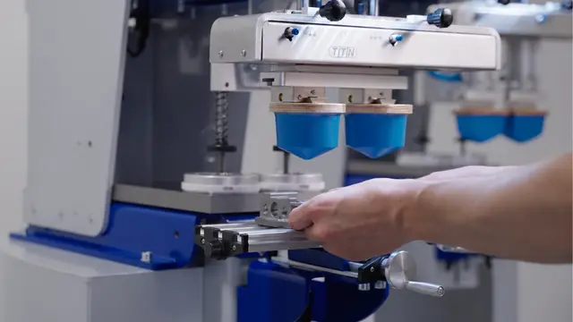 A close-up of a Universal Series pad printer from Automark, with a human hand carefully positioning a small, intricate aerospace part for precise marking, showcasing advanced pad printing technology for critical components