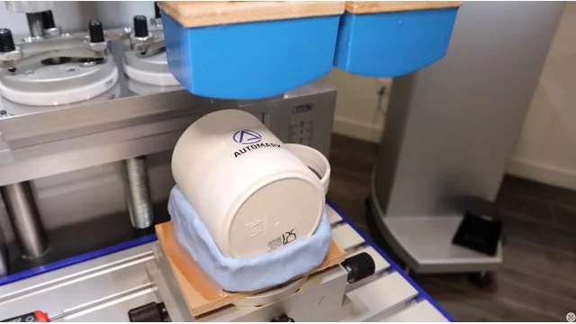 Close-up of a pad printer performing multi-color registration on a white coffee cup with the Automark logo, demonstrating precise alignment for complex designs.